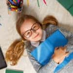 Smiling little girl in glasses lying on the floor with books, art supplies, and a tablet