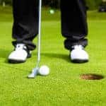 low shot of golfer's shoes, putter, and golf ball by the hole of a green