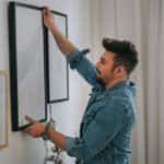 a middle eastern man with beard hanging a painting on the wall at his living room