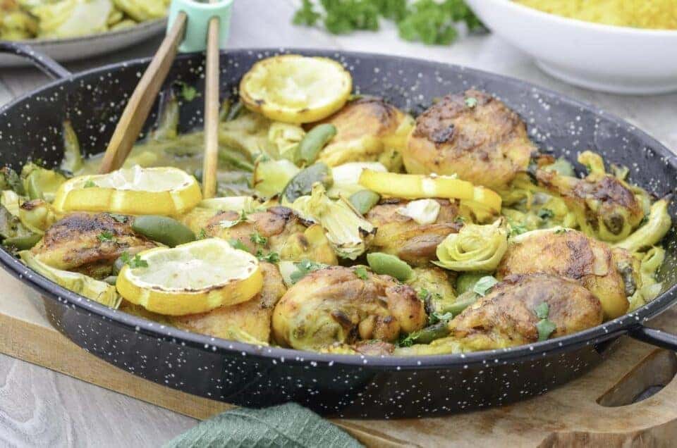 Crispy baked chicken thighs and drumsticks stew with artichokes hearts, green olives, roasted lemon slices, seasoning and onions served in a black enamelled pan on a wooden cutting board