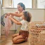 Young hispanic mother and her daughter sorting dirty laundry in the washing machine at home. Adorable little girl and her mother doing chores together at home