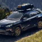 Toyota RAV4 with camping gear on top. The RAV4 is driving on a hill or mountain with a dirt road and surrounded by pine trees