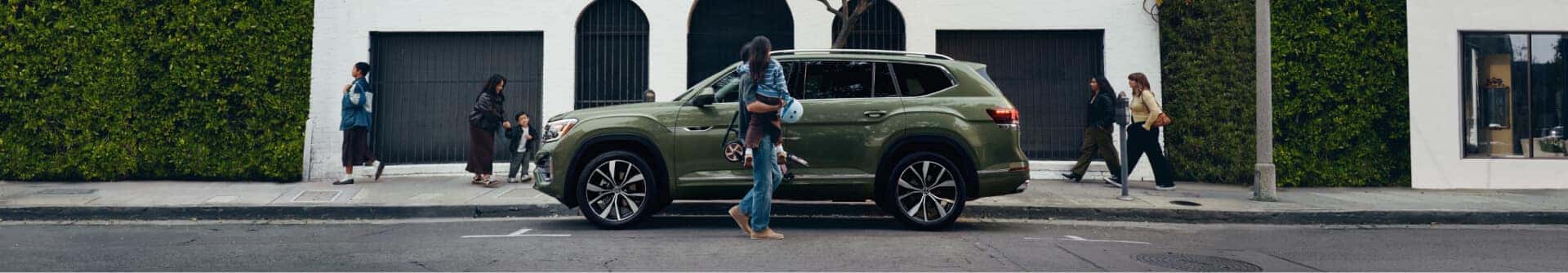 Profile view of a Volkswagen Atlas in Avocado Green as a family walks by on an urban city.