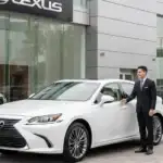 A Lexus dealership associate hands keys to a customer standing beside a white Lexus sedan outside a modern Lexus showroom, representing Certified Pre-Owned Lexus service and warranty coverage at Lexus of Mobile.