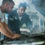 Auto mechanics cooperating while examining overheated car engine in auto repair shop. Focus is on man in blue uniform.