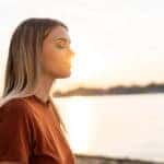 Young woman meditating on the riverside, enjoying the last sun rays of the day