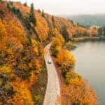 Car driving down a winding fall road near a lake.