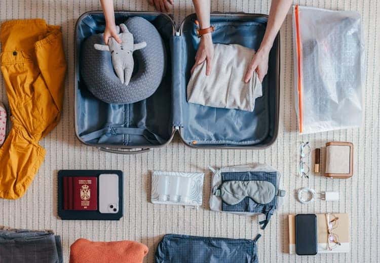 Flat lay of an anonymous family packing various things and essentials for their upcoming trip or vacation. Unrecognizable little girl putting in a toy in the suitcase. There is passport, mobile phone, travel pillow and other things laid on the carpet on the floor.