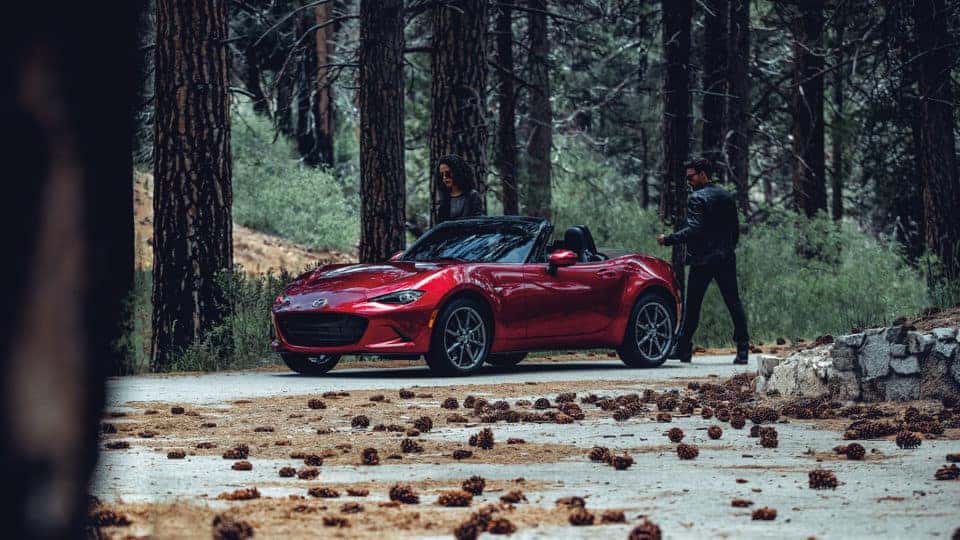 A red 2020 Mazda Miata in a forest; a couple is about to enter the vehicle.