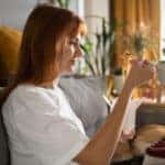 Side view of glad female eating Chinese food while sitting on couch near dog in cozy living room